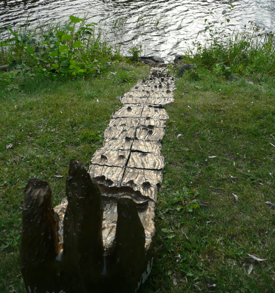 Auringosta virtaa / Streams from the sun. Kultalasitteinen keraaminen veistos Vanajaveden rannalla Hämeenlinnassa. Ceramic sculpture resembling a small golden stream by waterfront.