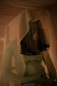 Green fabric sculpture sitting on a chair, wearing white and black veils.