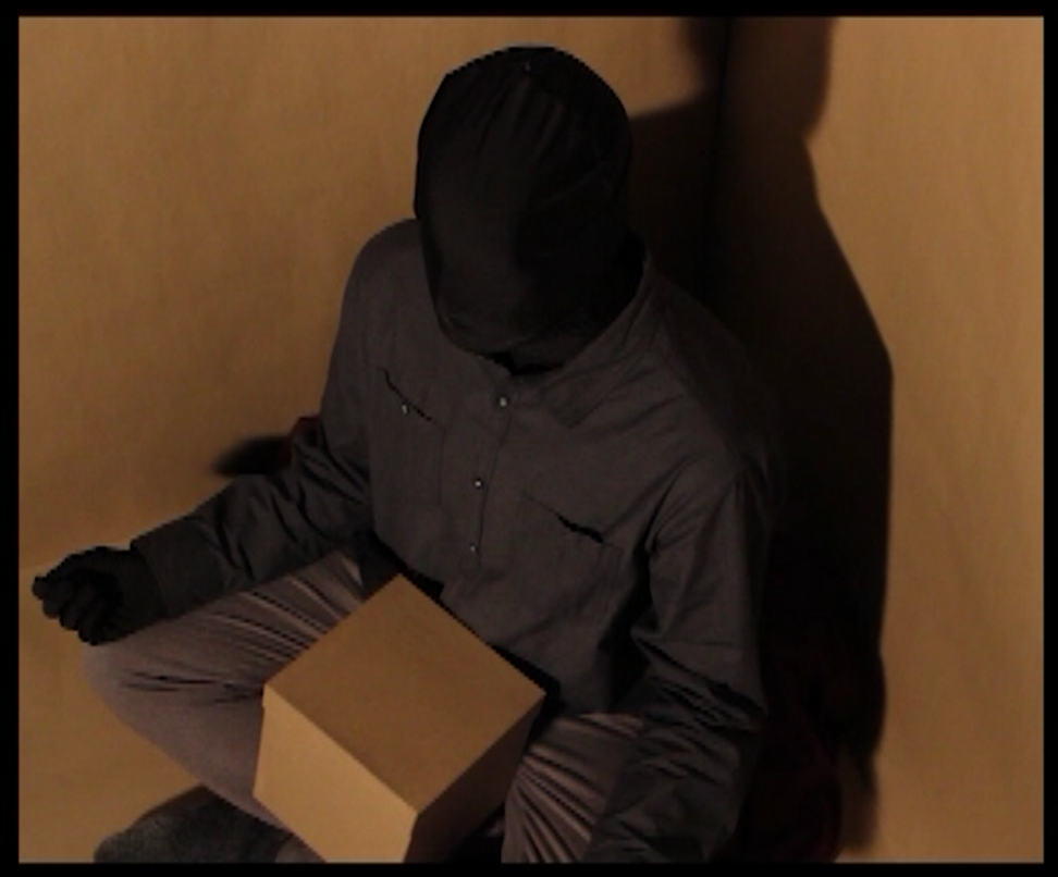 A darkened figure sits in a box with a box in their lap.